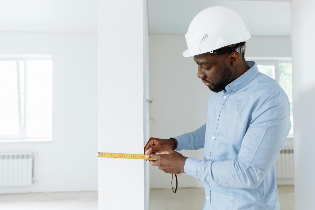 Construction worker measuring a wall indoors, wearing a white hard hat and blue shirt.